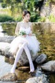 A woman in a white dress sitting on a rock by the water.